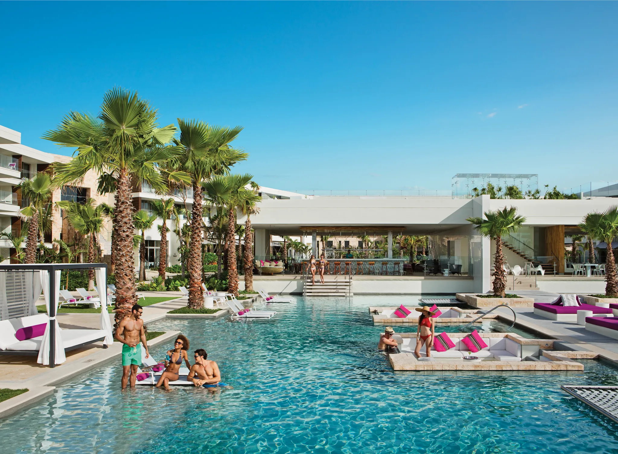 Guests enjoying the Energy Pool and The Bar area at Breathless Riviera Cancun, surrounded by palm trees and modern architecture.